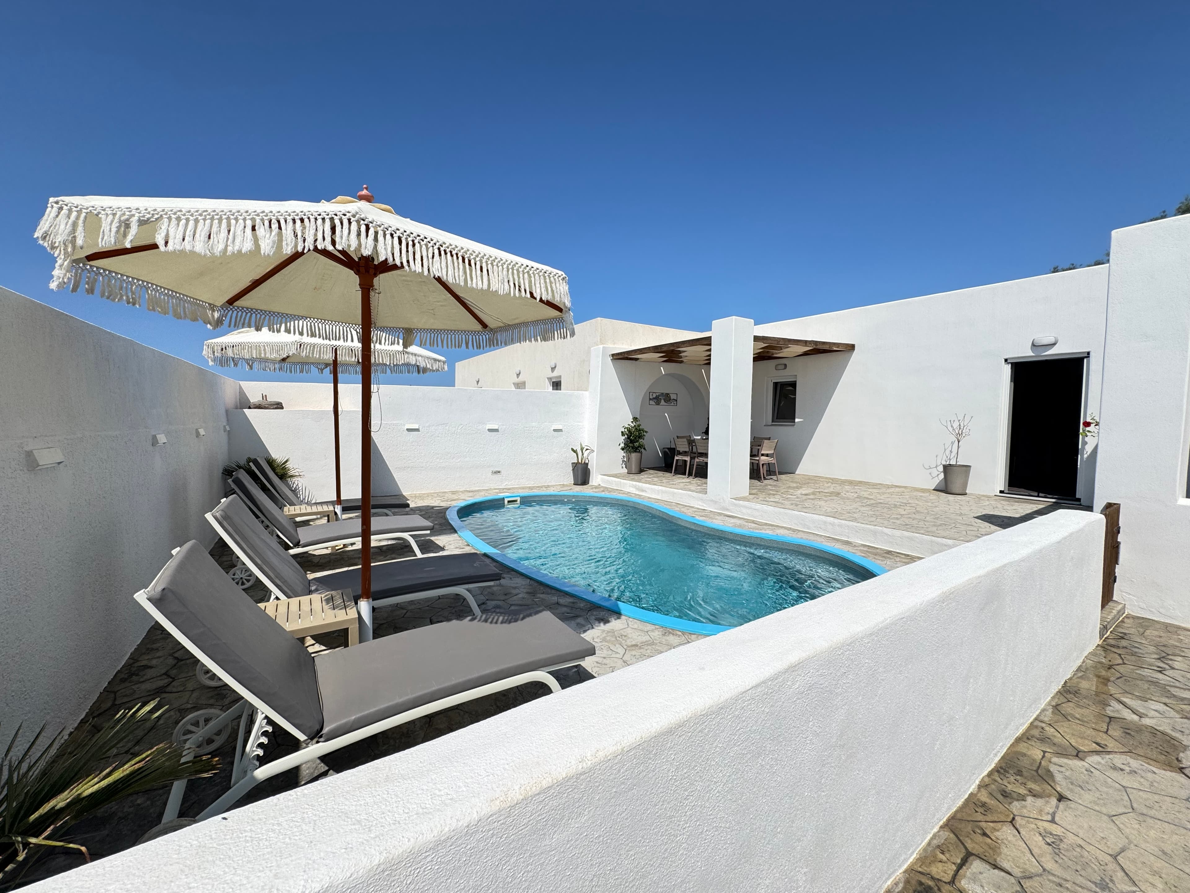 Small blue pool with gray lounge chairs and a white fringed umbrella in a courtyard.