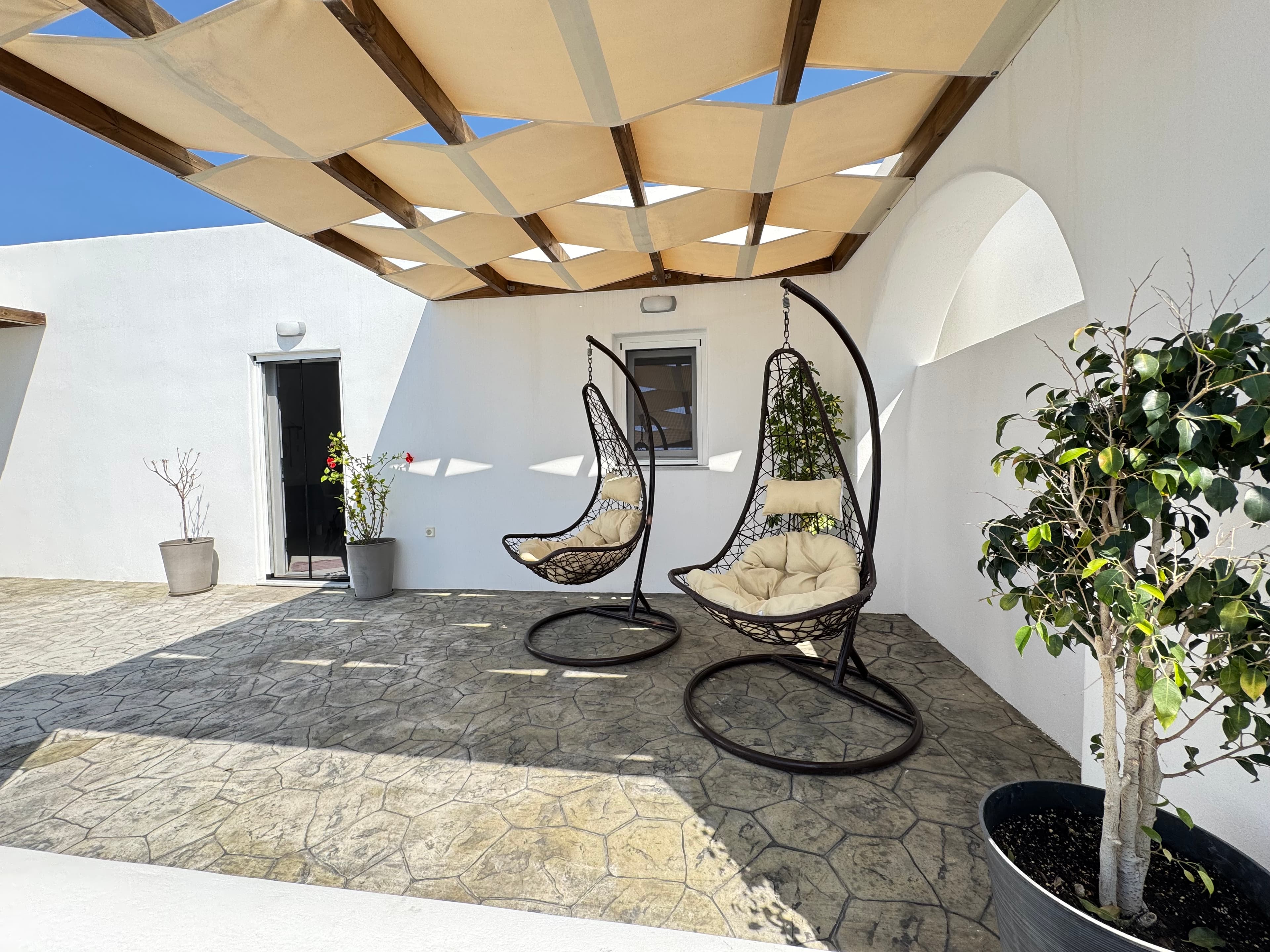 Two hanging egg chairs on a stone patio under a shaded pergola with white walls.