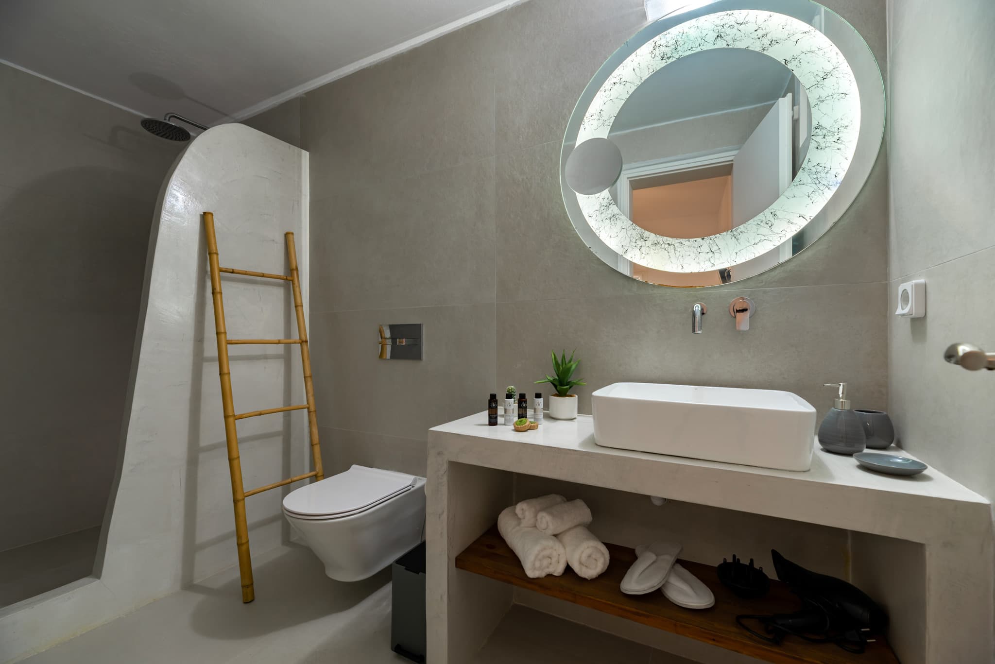 Modern grey bathroom with a round backlit mirror, white vessel sink, and bamboo ladder.