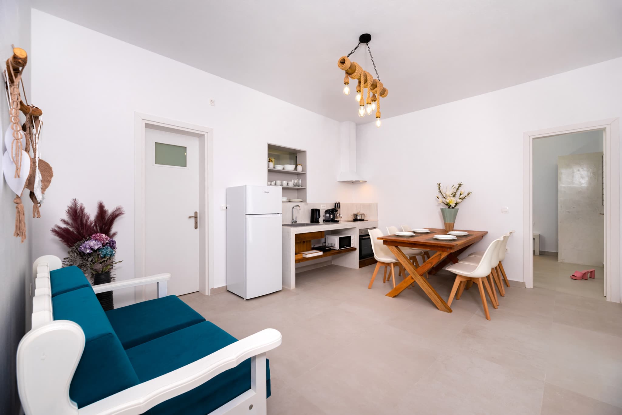 Modern white apartment interior with teal sofa, kitchenette, and wooden dining table with white chairs.