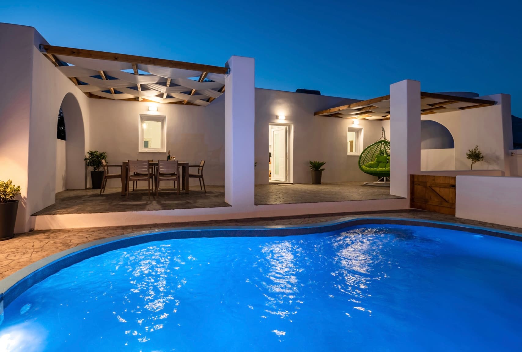 Modern white villa at dusk with a glowing blue swimming pool and outdoor patio.